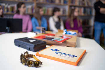 Verschillende soorten gegevensdragers op tafel met op de achtergrond scholieren die aandachtig zitten te luisteren naar de archivaris van het VRT Archief