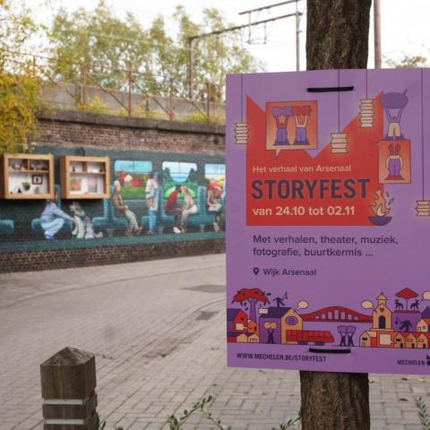 Op de voorgrond een reclamebord van Storyfest op de achtergrond een muurschildering en vitrinekasten met kunst gemaakt door bewoners uit de wijk Arsenaal
