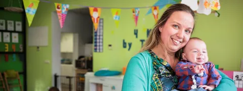 Een kinderopvangster houdt een baby vast terwijl er achter haar kindjes aan het spelen zijn.